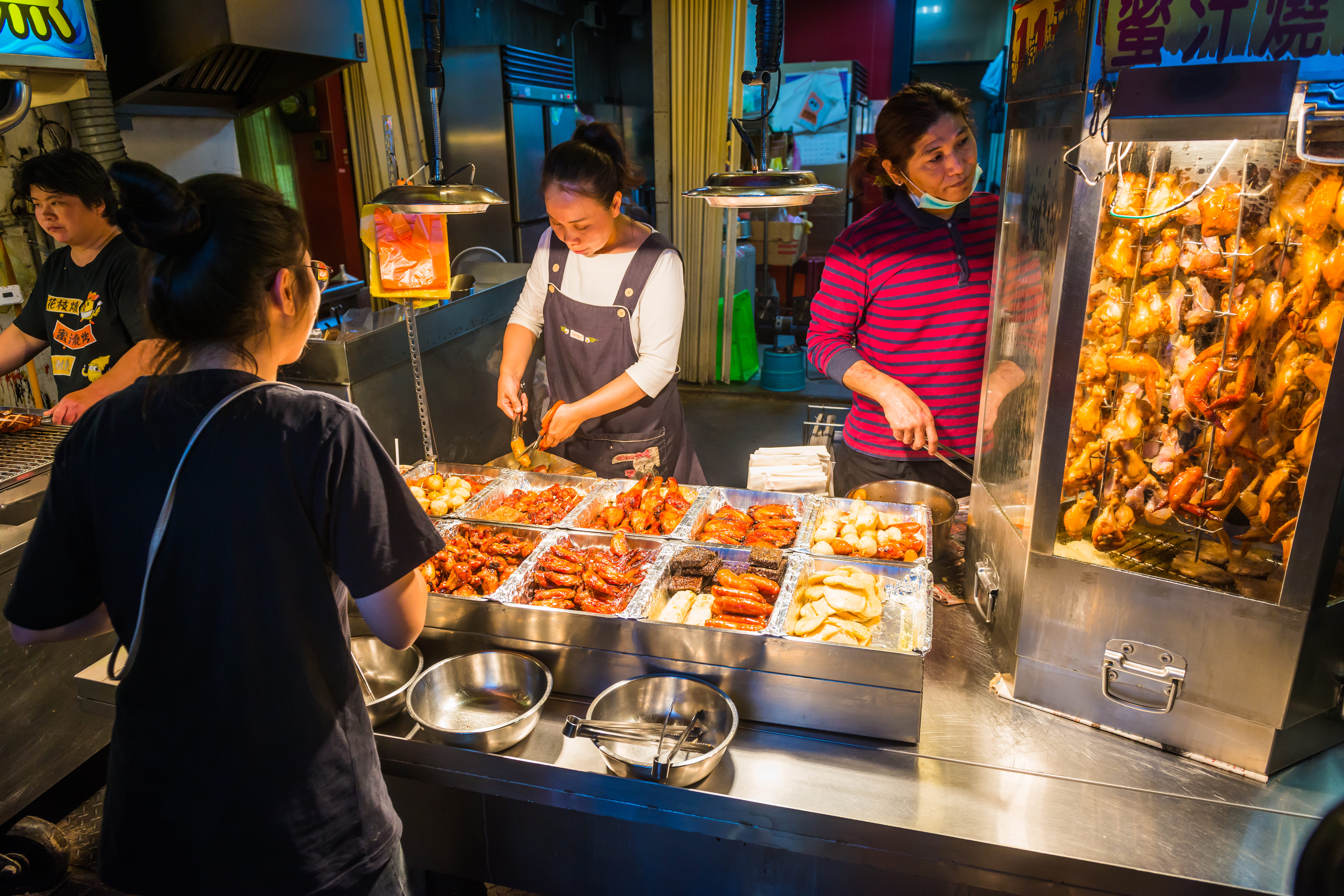 台灣夜市小吃攤位，店員製作傳統美食，展示多種油炸烤製小吃，顧客排隊購買，玻璃櫃內掛滿雞肉串，熱鬧繁忙的街頭美食文化場景