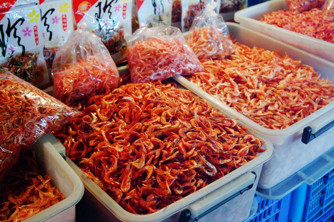 魚市場攤位上,白色大方籃內裝滿了成堆的橘紅色乾蝦米,後面還有幾袋包裝好的