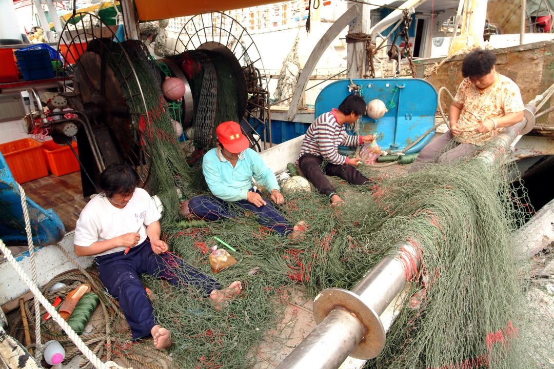 四名漁工坐在漁船甲板上,專注地用手修補綠色的漁網