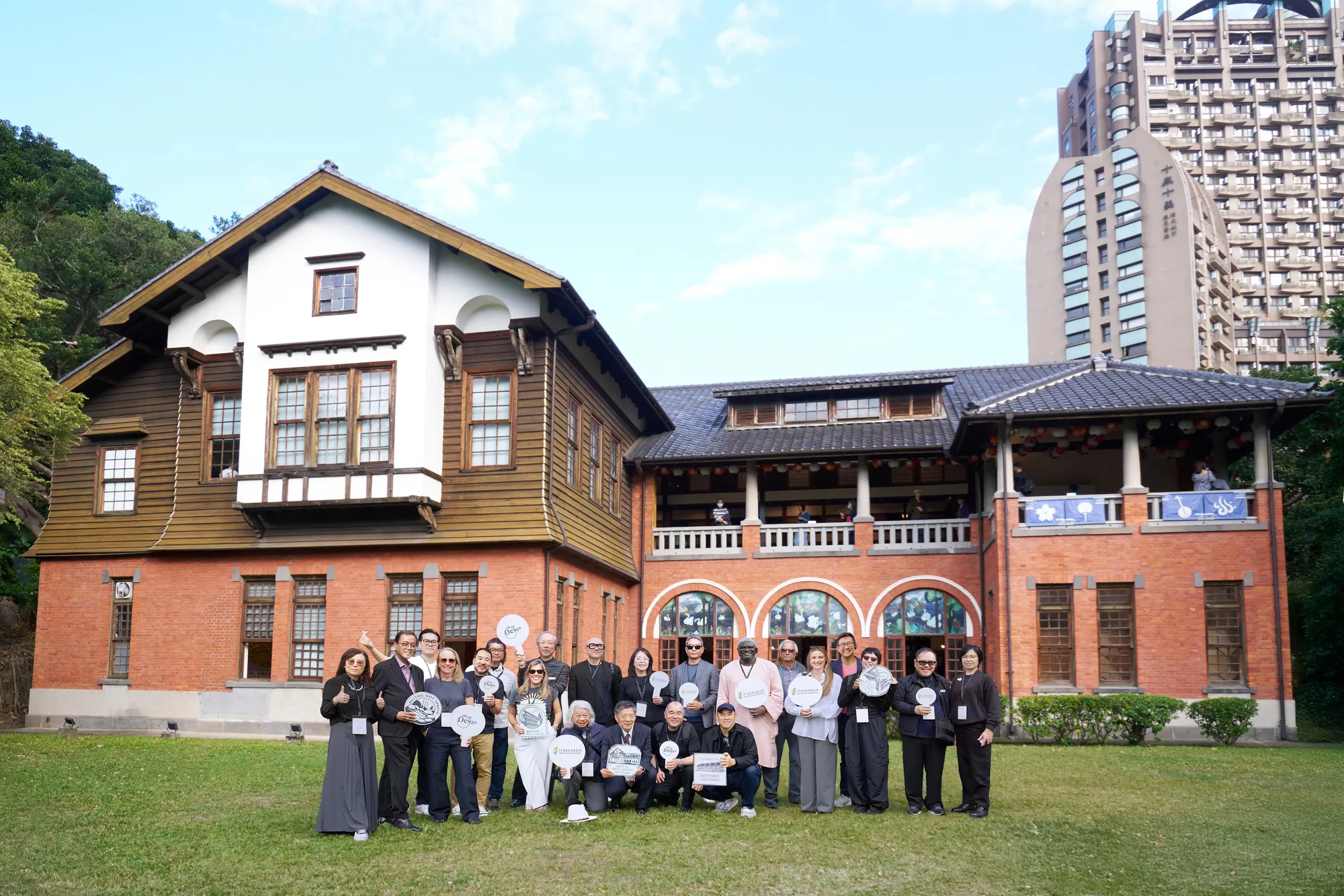 來自世界各地的設計師與貴賓在北投溫泉博物館紅磚建築前的大合照，眾人手持「看見台灣基金會」手拿牌，在藍天綠地中展現跨國文化交流的活力。